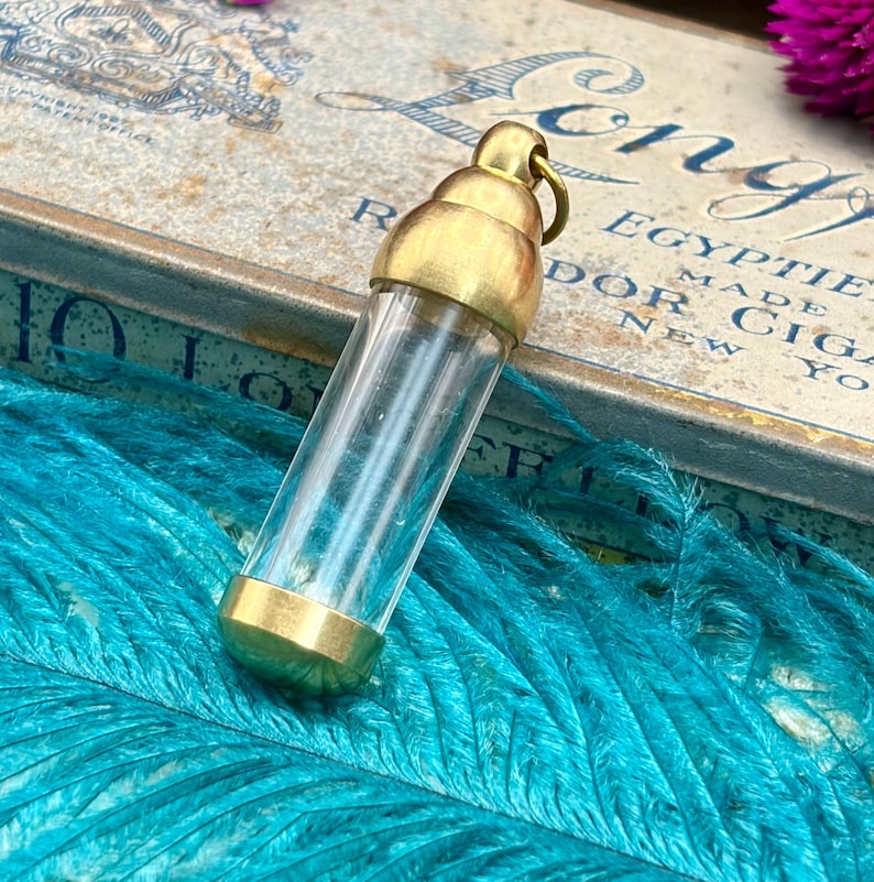 May include: A clear glass vial with brass-coloured metal caps and a loop at the top. The vial is cylindrical and sits on a bed of turquoise feathers. The background includes a vintage box with text.