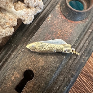 Puede incluir: Un cuchillo de bolsillo de color dorado en forma de pez con una hoja de plata. El cuchillo está sobre una superficie de madera marrón oscuro con un agujero de cerradura.