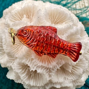 May include: A red and orange fish-shaped pendant with a gold-colored loop. The pendant has detailed scales and fins. The pendant is resting on a white, textured surface.