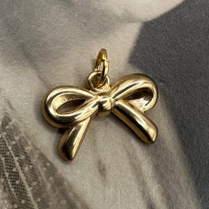 May include: A gold-colored bow charm pendant. The bow is tied in a knot and has a smooth, shiny finish.