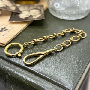 Puede incluir: Una cadena dorada con un cierre y un anillo. La cadena está hecha de eslabones retorcidos y en bucle. El cierre es de resorte. La cadena descansa sobre un libro verde oscuro.