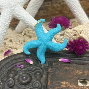 May include: A turquoise starfish pendant with a gold-colored ring. The starfish is set against a backdrop of white starfish, purple flowers, and a cream-colored crocheted fabric. The pendant is a vibrant accent.