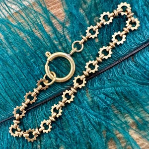 May include: A gold-colored bracelet with a unique design featuring a series of interconnected, gear-like shapes. The bracelet has a circular clasp and is displayed against a backdrop of teal feathers.