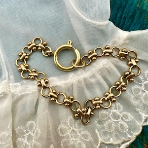 May include: A gold-toned bracelet with a circular clasp. The bracelet features a series of small, connected circles and decorative floral-like elements. The jewelry is displayed on a white lace surface.