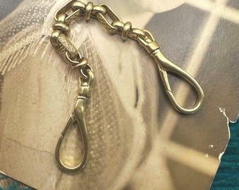 May include: A vintage gold-toned watch chain with two clasp ends and a decorative chain link design. The chain is displayed on a sepia-toned photograph, showcasing its antique aesthetic. The chain is likely made of brass or a similar metal.