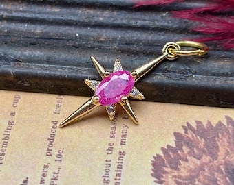 May include: A gold-colored pendant in the shape of a starburst, featuring a large, oval-shaped pink gemstone at its center. The starburst has pointed rays and is accented with smaller, clear gemstones. The pendant is suspended from a small loop.