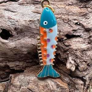 May include: A gold-colored fish charm with a teal, white, orange, and purple enamel design. The fish has a white body with orange and purple polka dots, a teal head and tail, and a gold loop for hanging.