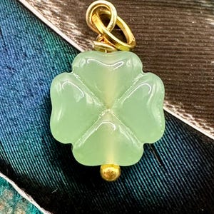 May include: A light green, four-leaf clover pendant with a gold-colored bail and a small gold bead at the bottom. The pendant is made of a translucent material and is set against a background of blue and green fabric.