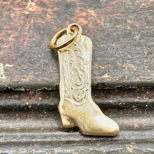 Puede incluir: Encanto de metal dorado en forma de bota vaquera con un diseño detallado. La bota tiene una punta puntiaguda y un pequeño lazo en la parte superior para sujetarlo a un collar o pulsera.