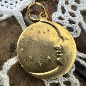 May include: A gold-toned crescent moon charm with a face and stars. The charm is on a white crocheted surface.