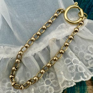 May include: A gold-toned bracelet featuring a chain design with alternating small beads and oval links. The bracelet has a large, round clasp. The jewelry is displayed on a white, lacy fabric.