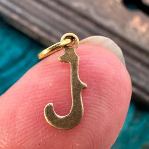 May include: Gold-toned metal charm in the shape of the letter J. The charm has a decorative, ornate design.