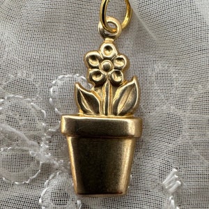 May include: A gold-coloured charm in the shape of a potted flower. The flower has five petals and a centre circle. The pot is a simple, rounded shape.