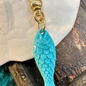 May include: A turquoise blue fish charm with a gold chain and clasp. The fish has a detailed design with scales and a fin.