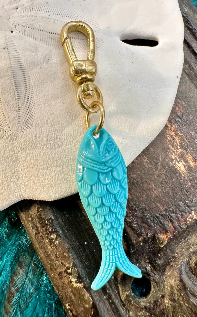 May include: A turquoise blue fish charm with a gold chain and clasp. The fish has a detailed design with scales and a fin.