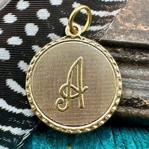 May include: Gold-toned pendant charm featuring a raised, cursive letter 'A' on a round, textured surface. The pendant has a loop at the top for attaching to a chain or bracelet.