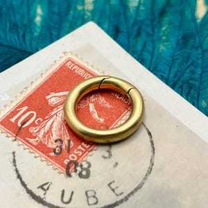 May include: A gold-colored, circular clasp is the focal point, set against a vintage postage stamp with red and white hues. The stamp features a woman's portrait and text. The clasp is open, revealing its hinge.