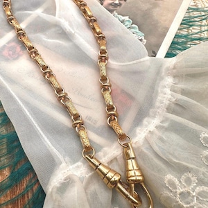 Puede incluir: Un collar dorado con un diseño de cadena decorativa y dos extremos de cierre. El collar se muestra sobre una tela blanca y con encaje, con una postal vintage y un guante blanco en el fondo.