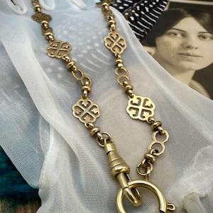 May include: A gold-toned necklace with a decorative clasp and ornate links. The necklace features repeating cross-like designs and is displayed on a white, sheer fabric. A vintage photograph and a feather are in the background.