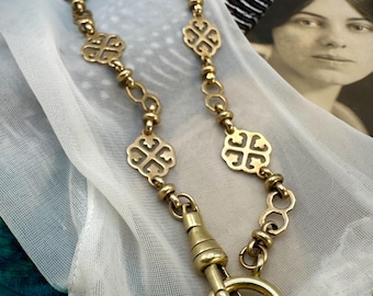 Puede incluir: Un collar dorado con un cierre decorativo y eslabones ornamentados. El collar presenta diseños repetidos en forma de cruz y se muestra sobre una tela blanca y transparente. Una fotografía vintage y una pluma están en el fondo.