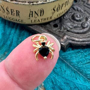 Puede incluir: Un dije de araña dorado con un cuerpo negro en forma de corazón y ocho patas. El dije es pequeño y detallado, con un lazo en la parte superior para sujetarlo a un collar o pulsera. El dije está sostenido por un dedo.