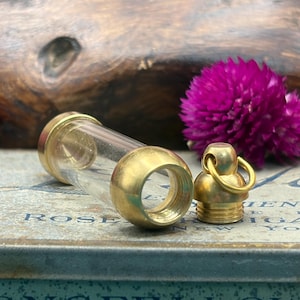 May include: A clear glass vial with brass-coloured metal ends and a matching screw-on cap. The vial is lying on a surface with the word "ROSE" visible. A vibrant purple flower is in the background.