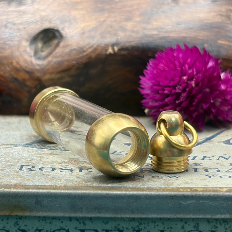 May include: A clear glass vial with brass-coloured metal ends and a matching screw-on cap. The vial is lying on a surface with the word "ROSE" visible. A vibrant purple flower is in the background.