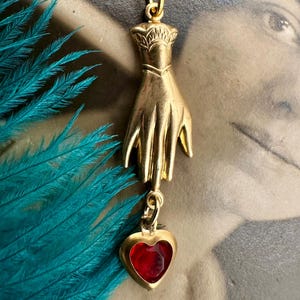 Puede incluir: Un colgante de oro en forma de mano sosteniendo un dije de corazón rojo. La mano está detallada con una superficie texturizada y el corazón tiene un centro de vidrio rojo.