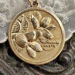 May include: A gold-toned pendant featuring a raised design of two violet flowers with the text "February Violets" inscribed on the front.