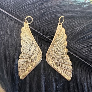 May include: A pair of gold-tone metal angel wing charms with detailed feather designs. The charms are on a black feather background.