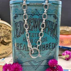 May include: A silver-toned chain necklace featuring elongated oval links and a circular spring-ring clasp. The necklace is draped over a rectangular, light blue metal tin with black text that reads EDGEWORTH EXTRA HIGH GRADE READY RUBBED.