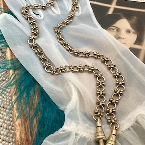 Könnte beinhalten: Eine goldfarbene Vintage-Halskette mit einem einzigartigen Kettendesign, das abwechselnd Glieder und kleine Kugelakzente aufweist. Die Halskette hat unten zwei Verschlüsse. Die Halskette wird auf einem weißen Handschuh mit einem Vintage-Foto im Hintergrund präsentiert.
