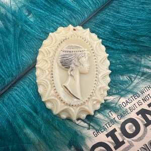 May include: A white cameo brooch with a detailed design of a woman's profile. The cameo is surrounded by a decorative border with small beads.