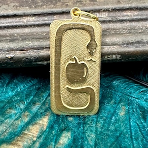 May include: A rectangular gold-colored pendant featuring a snake coiled around an apple. The snake's head is near the top, and the apple is in the center. The pendant has a textured background and a gold-colored loop at the top for hanging.