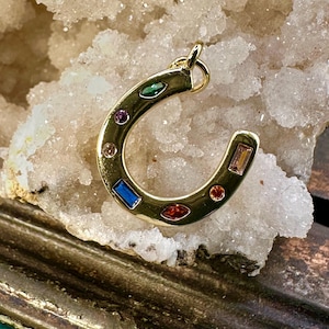 May include: A gold-colored horseshoe pendant adorned with various colorful gemstones. The gemstones include blue, red, green, and pink, set within the curved shape of the horseshoe. The pendant is designed for jewelry.