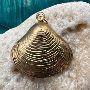 May include: A gold-colored clam shell pendant with a detailed textured surface. The pendant is on a white background.