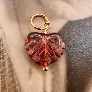 May include: A gold-colored metal charm with a leaf-shaped, reddish-brown glass pendant. The pendant has a detailed leaf vein design and a small gold bead at the bottom. The charm is designed to be attached to a necklace or bracelet.
