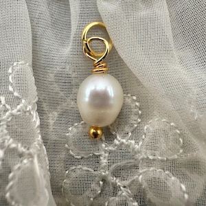 Puede incluir: Un charm dorado con una perla blanca y un bucle dorado. El charm está sobre un fondo de encaje blanco.