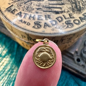 May include: Gold-toned charm featuring a detailed crab design. The charm is round and has a loop at the top for attaching to a necklace or bracelet.