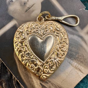 May include: A gold heart-shaped locket with intricate floral designs and a smaller heart in the center. The locket has a clasp on the top for attaching to a chain or keychain.