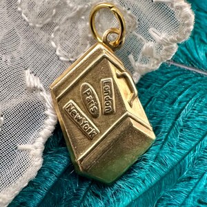 May include: Gold-toned metal charm shaped like a suitcase with the words "London", "Paris", and "New York" embossed on the sides.