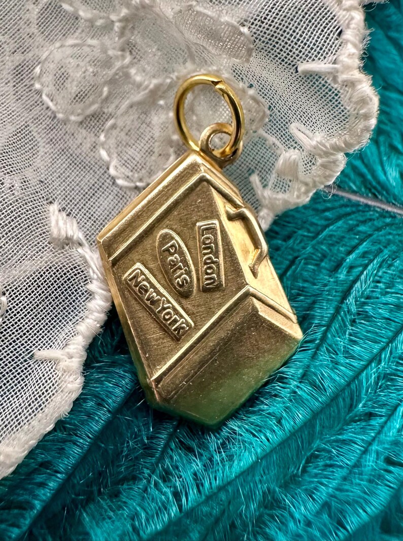 May include: Gold-toned metal charm shaped like a suitcase with the words "London", "Paris", and "New York" embossed on the sides.