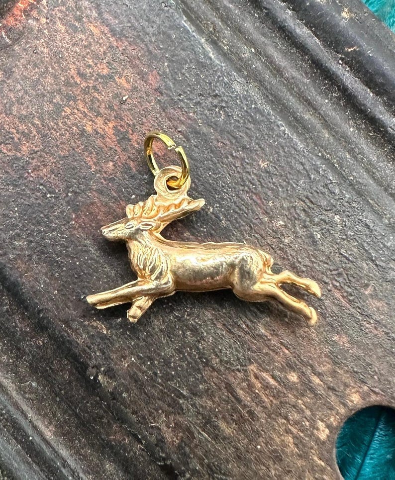 K&ouml;nnte beinhalten: Ein goldfarbener Hirsch-Anh&auml;nger. Der Hirsch ist in einer laufenden Pose dargestellt, mit dem Kopf zur Seite gedreht. Der Anh&auml;nger hat eine Schlaufe oben zum Anbringen an einer Halskette oder einem Armband.