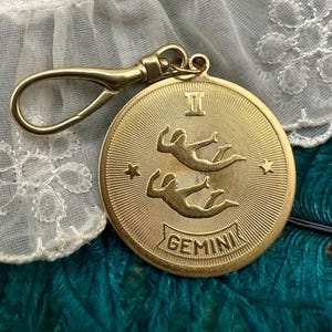 May include: A gold-coloured Gemini zodiac charm with a clasp. The charm features two figures, stars, and the word "Gemini". The charm is round with a textured background.