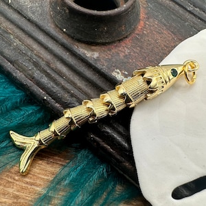 May include: A gold-tone fish charm with a green gemstone eye. The fish is made of metal and has a detailed design.