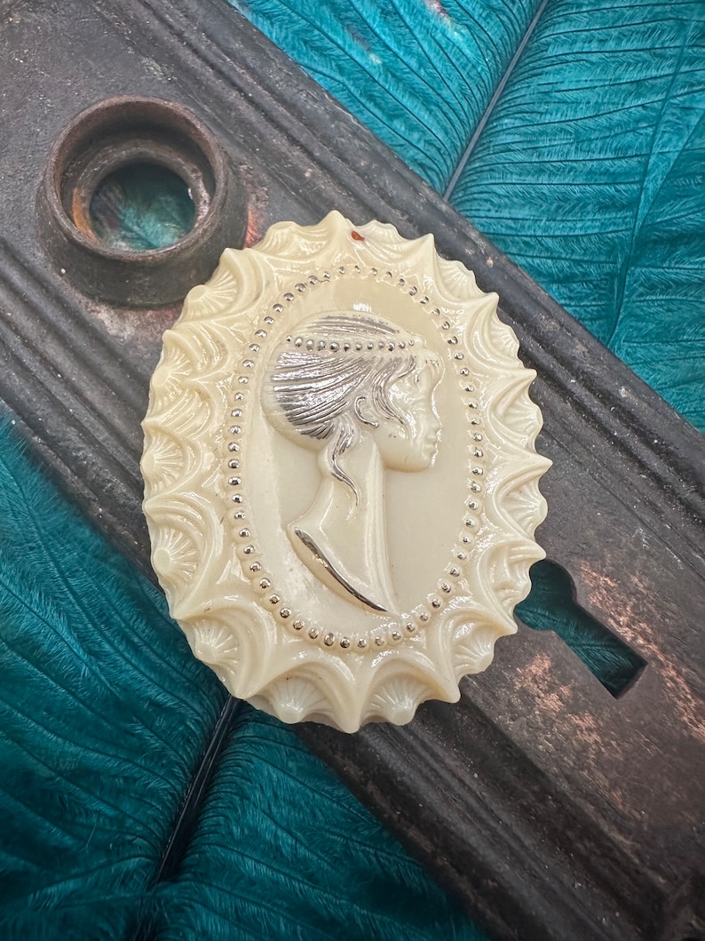 May include: A white cameo brooch with a silver-toned design featuring a woman's profile. The cameo is oval-shaped and has a decorative border.