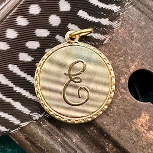 May include: A gold-coloured pendant with a circular design featuring a raised, stylised letter 'E' in the centre. The pendant has a textured surface and a beaded edge.