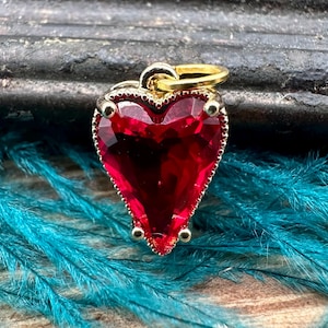 May include: A heart-shaped pendant with a faceted, deep red gemstone. The pendant is framed in gold-colored metal with small, round accents and a loop for a chain. The background features a dark surface and teal feathers.