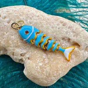 May include: A colorful fish-shaped pendant with a gold-tone bail. The pendant features a light blue body with yellow and white accents. A small, orange gem is set in the eye. The fish is resting on a light-colored stone.