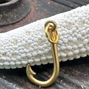May include: A gold-colored metal fishing hook charm with a loop at the top. The hook is on a white textured surface.